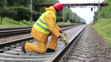 Demiryolu işçisi sarı üniformalı ve demiryolu hattında ölçüm aleti olan. Demiryolu işçisi kırmızı şapkalı tren raylarında oturur ve trene bakar. Seviye ölçüm aletine sahip işçiler