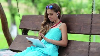 Çekici kız salıncakta oturuyor ve tablet bilgisayarı tutuyor. Tabletli güzel kız bahçede bankta oturuyor. Tablet bilgisayarında oyun oynayan bir kız. Tabletli güzel kız.