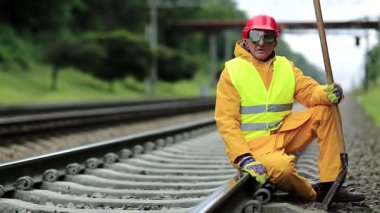 Sarı üniformalı, elinde kürekle demiryolu işçisi demiryolu hattında oturuyor. Demiryolu işçisi kırmızı başlıklı adam raylara oturur ve kameraya bakar. Demiryolunda kürekli bir işçi. Demiryolu inşaatı
