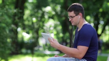Adam şehir parkındaki bankta oturuyor ve tablet bilgisayarla iletişim kuruyor. Mavi tişörtlü yetişkin adam halka açık bir bahçede bankta oturuyor ve tablet bilgisayarında konuşuyor. Tabletli adam Skype üzerinden iletişim kuruyor.