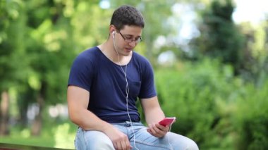 Adam şehir parkında bankta oturuyor ve akıllı telefondan konuşuyor. Adam akıllı telefonla iletişim kuruyor. Adam halka açık bir bahçede bankta oturuyor ve Skype üzerinden iletişim kuruyor.
