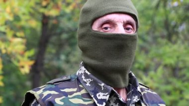 Askeri üniformalı ve maskeli bir adam elinde siyah bir tabanca olan kameraya bakıyor. Asker hedefe nişan alır. Poligondaki emekli memur. Ormanda askeri üniformalı kıdemli bir adam. Silahlı adam.