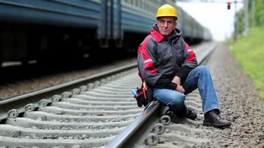 Demiryolu işçisi demiryolu hattında oturur. Demiryolunda tren yolculukları. Sarı şapkalı demiryolu işçisi tırabzanda oturur ve kameraya bakar. Demiryolu işçisi
