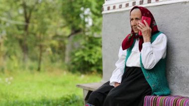 Son sınıf öğrencisi kadın bankta oturuyor ve cep telefonuyla konuşuyor. Yaşlı kadın evinin yanındaki bankta oturuyor ve cep telefonuyla konuşuyor. Kırmızı akıllı telefonlu Ukraynalı yaşlı kadın. Akıllı telefonlu bir kadın. Kahkaha
