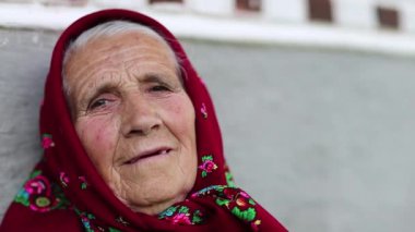 Kıdemli kadın kameraya bakar ve gülümser. Kırmızı başörtülü yaşlı kadın evinin gri duvarının yanında oturuyor ve kameraya bakıyor. Kadın kameraya bakıyor ve gülümsüyor, buruşuk surat.