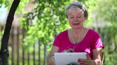 Yaşlı kadın bahçede sandalyede oturuyor ve tablet bilgisayarla iletişim kuruyor. Tablet bilgisayarı olan bir kadın. Tabletli kadın bahçede ağacın altında oturuyor. Tabletli kadın Skype üzerinden iletişim kuruyor.