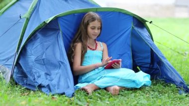 Güzel kız turist çadırının yanındaki çimlere oturur ve akıllı telefon kullanır. Kırmızı akıllı telefonlu açık mavi elbiseli kız çimlerin üzerinde oturuyor. Güzel kız onun akıllı telefonundaki fotoğraflara bakar ve bakar.