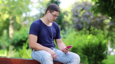 Genç adam şehir parkındaki bankta oturuyor ve akıllı telefondan konuşuyor. Adam halka açık bir bahçede bankta oturuyor ve cep telefonu kullanıyor. Adam akıllı telefonla iletişim kuruyor.