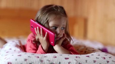 Akıllı telefonlu komik kız yatakta yatıyor. Küçük kız yastığa uzanmış, elini tutuyor kırmızı cep telefonu ve el kol hareketi yapıyor.