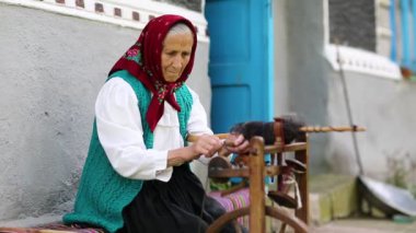 Yaşlı kadın bankta oturuyor ve dönen tekerlekle distaff ile çalışıyor. Ukraynalı yaşlı kadın evinin yanındaki bankta oturur ve iplik örer. Dönen tekerli bir kadın, el işi.