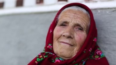 Yaşlı kadın kameraya bakıyor. Kırmızı başörtülü yaşlı kadın evinin gri duvarının yanında oturuyor ve kameraya bakıyor. Kadın kameraya bakıyor ve gülümsüyor, buruşuk surat.