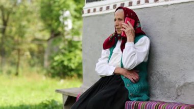 Son sınıf öğrencisi kadın bankta oturuyor ve cep telefonuyla konuşuyor. Yaşlı kadın evinin yanındaki bankta oturuyor ve cep telefonuyla konuşuyor. Kırmızı akıllı telefonlu Ukraynalı yaşlı kadın. Akıllı telefonu olan bir kadın.