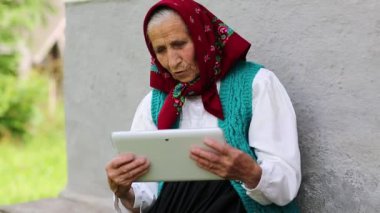 Yaşlı kadın bankta oturuyor ve tablet bilgisayarla iletişim kuruyor. Yaşlı kadın evinin yanındaki bankta oturuyor ve Skype 'ta tablet bilgisayarla konuşuyor. Beyaz tabletli Ukraynalı yaşlı kadın. Tablet pc ile kadın