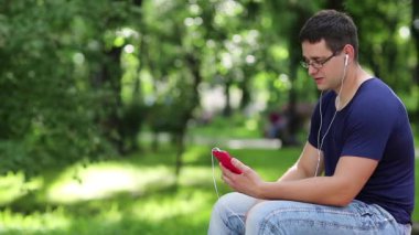 Genç adam şehir parkındaki bankta oturuyor ve akıllı telefondan konuşuyor. Adam kırmızı akıllı telefonla iletişim kuruyor. Adam halka açık bir bahçede bankta oturuyor ve cep telefonuyla Skype üzerinden iletişim kuruyor.