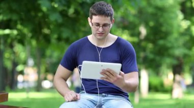 Genç adam şehir parkındaki bankta oturuyor ve tablet bilgisayarla iletişim kuruyor. Mavi tişörtlü adam halka açık bir bahçede bankta oturuyor ve tablet bilgisayarında konuşuyor. Tabletli adam Skype üzerinden iletişim kuruyor.