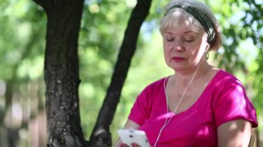 Sarışın kadın bahçede oturur ve akıllı telefon aracılığıyla iletişim kurar. Beyaz akıllı telefonlu bir kadın. Akıllı telefonlu iş kadını bahçedeki ağacın altında oturuyor. Kadın cep telefonuyla Skype üzerinden konuşuyor.