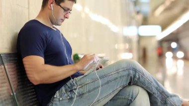 Tablet bilgisayarı olan adam yeraltı istasyonundaki bankta oturuyor. Mavi tişörtlü yetişkin adam metro istasyonundaki metal bankta oturuyor ve tablet bilgisayar kullanıyor. Tablet bilgisayarlı bir iş adamı