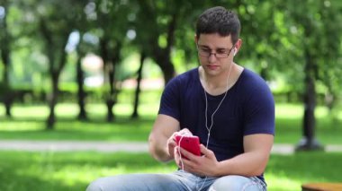 Kırmızı akıllı telefonlu yetişkin bir adam halka açık bir bahçede bankta oturuyor. Mavi tişörtlü adam şehir parkında bankta oturuyor ve cep telefonu kullanıyor. Adam kulaklıklı kırmızı kişisel cep telefonu kullanıyor.