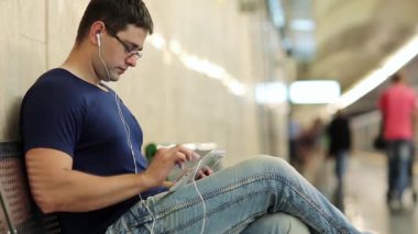 Tabletli yetişkin adam yeraltı istasyonundaki bankta oturuyor. Adam metro istasyonundaki metal bankta oturuyor ve tablet bilgisayar kullanıyor. Tablet bilgisayarlı bir iş adamı. Tablet bilgisayarlı erkek