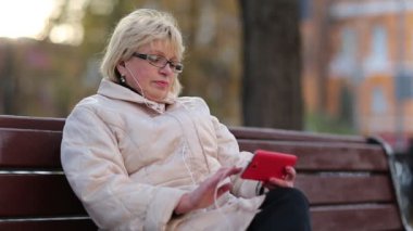 Kadın yolun yanındaki bankta oturuyor ve kırmızı akıllı telefon kullanıyor. Sarışın kadın akıllı telefondan fotoğraflara bakıp göz gezdiriyor. Akıllı telefonlu bir kadın. Cep telefonu olan bir iş kadını