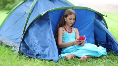 Güzel kız mavi turist çadırının yanındaki çimlerde oturur ve akıllı telefon aracılığıyla iletişim kurar. Açık mavi elbiseli, kırmızı akıllı telefonlu güzel bir kız çimlerin üzerinde oturuyor. Güzel kız Skype üzerinden telefonda konuşuyor.