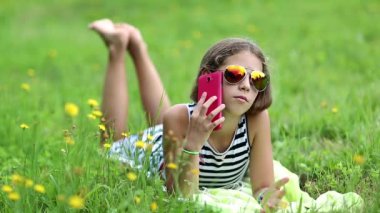 Kırmızı akıllı telefonlu güneş gözlüklü güzel kız çimlerin üzerinde yatıyor. Gözlüklü çekici kız yeşil çimenlere uzanıyor ve cep telefonu kullanıyor. Akıllı telefonu olan bir kadın.