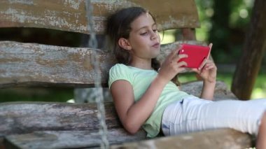 Kırmızı akıllı telefonlu bir kız bahçede salıncakta oturuyor ve videoya bakıyor. Çekici kız akıllı telefon kullanıyor. Kırmızı akıllı telefonlu güzel kız salıncakta oturuyor.