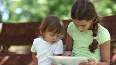 Tabletli çocuklar. Beyaz tabletli iki küçük kız salıncakta oturuyor. Tablet bilgisayarı olan bir kadın. Tabletli iki kız kardeş bahçede salıncakta oturuyor.