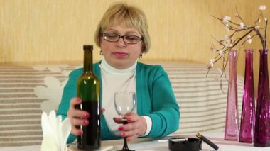 Elinde bir şişe şarap olan yaşlı bir kadın masada oturuyor. Kadın şarap içiyor.