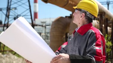 Isı elektrik santralindeki ana ısıtma hattının yanında mühendislik çizimleri olan sarı şapkalı bir işçi. Isı elektrik santralinde proje çizimleri yapan bakım mühendisi