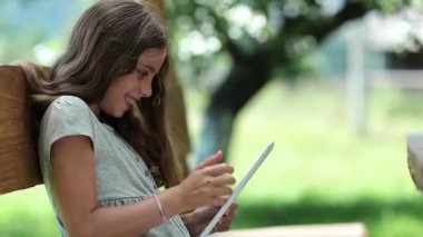 Beyaz tablet bilgisayar ile güzel kız oyunu oynar. Kız onun Tablet Pc tablet bilgisayar ile küçük kız bahçede bankta oturur. oyunu oynar. Kadın, tablet bilgisayar