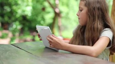 Güzel kız beyaz tablet bilgisayar ile masada oturur. Kız oyunu oynar onun Tablet Pc tablet bilgisayar ile küçük kız bahçede bankta oturur.