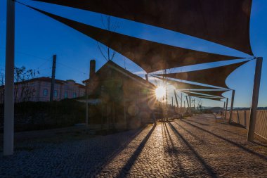 Gölgeli yelkenleri olan asfalt bir yol nehir boyunca akar, su manzarası ve uzak tepeler sunar.