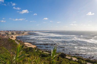 Sahile vuran dalgalar ve dağınık bulutlu mavi bir gökyüzü ile kayalık bir plaja yakın bir kasaba sahili.