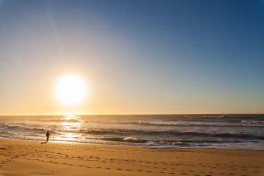 Günbatımında kumlu bir sahilde tek başına balık avlayan bir figür. Güneş okyanus ufkunun üzerinde batıyor..
