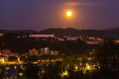 Aydınlık bir dolunay, şehri aydınlatan aydınlık binalar, dolambaçlı bir nehir ve çevreleyen tepelerle gece gökyüzünü aydınlatır..