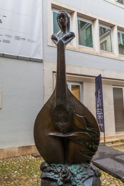 Portekiz gitarı çalan bir kadının bronz heykeli, fado enstrümanı olarak bilinir, arka planda kaldırım taşları ve bina cephesi olan bir avluda oturur..