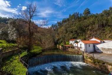 Şelalenin çağlayanı sıra kırmızı kiremitli çatıları olan geleneksel binalarla birlikte bir nehre aktığı, yemyeşil, parlak mavi bir gökyüzünün altında dağınık bulutlarla bezenmiş manzaralı bir manzara..