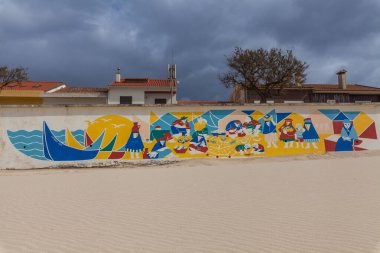 Moda figürleri ve tekneleri gösteren canlı bir duvar, kumlu bir alana yakın beyaz bir duvarı süslüyor. Arkaplanda binalar ve bulutlu bir gökyüzü var..