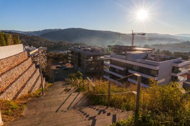 Çağdaş binaların, bir inşaat vincinin ve parlak, güneşli bir gökyüzünün altındaki uzak tepelerin yer aldığı bir taş merdiven modern bir şehir manzarasına iniyor.