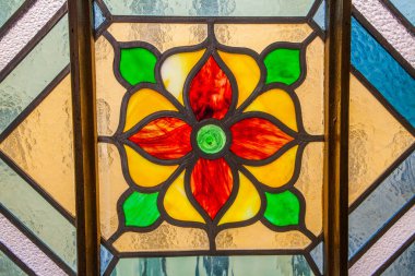 Rengi parlak kırmızı, sarı ve yeşil renkli merkezi çiçekli bir madalyonun sessiz tonlarda dokulu camlarla çevrili olduğu yakın plan bir fotoğraf. Kudüs sinagogu.