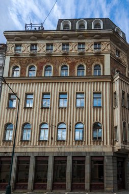 Süslemeli ön cephesi kemerli pencereler, zıt dokular ve bulutlu bir gökyüzünün altında koyu renkli üst katlı çok katlı bir bina.