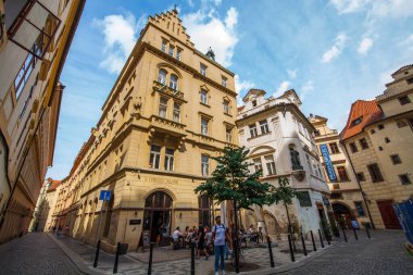 Çekici bir Avrupa sokak sahnesinde farklı mimari tarzlara sahip tarihi binalar, küçük bir ağaç, bir kafede müşterilerle birlikte açık havada oturma ve kaldırım taşı sokağının üzerinde parçalı bulutlu bir gökyüzü sergileniyor..