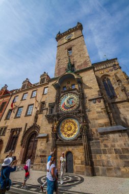 Avrupa 'da tarihi bir kilisenin bulunduğu geniş bir kaldırım taşı meydanı. Çarpıcı kuleler, renkli cepheli binalar, göze çarpan bir anıt ve parçalı bulutlu bir gökyüzü altında güneşli günün keyfini çıkaran çok sayıda insan..