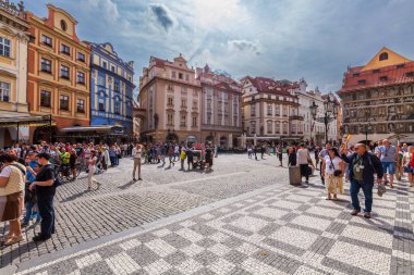 Avrupa şehrinde geniş bir kaldırım taşı meydanı. İçinde ikiz kuleleri olan tarihi bir kilise renkli cepheli binalar, önemli bir anıt ve parçalı bulutlu bir gökyüzü altında güneşli günün tadını çıkaran çok sayıda insan var..