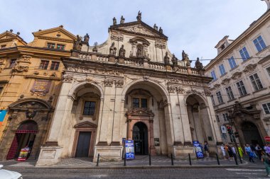 Heykellerin, kemerli açıkların ve dekoratif detayların yer aldığı simetrik cepheli bir barok tarzı kilise, bulutlu bir gökyüzünün altında etrafındaki binaların bulunduğu şehir meydanında belirgin bir şekilde duruyor..