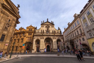 Simetrik cepheli, heykelleri, kemerli açıkları ve belirgin yeşil kubbesi olan barok tarzı bir kilise, bulutlu bir gökyüzünün altında çevresindeki binaların bulunduğu şehir meydanında belirgin bir şekilde duruyor.