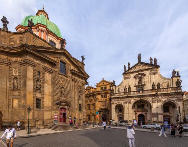 Simetrik cepheli, heykelleri, kemerli açıkları ve belirgin yeşil kubbesi olan barok tarzı bir kilise, bulutlu bir gökyüzünün altında çevresindeki binaların bulunduğu şehir meydanında belirgin bir şekilde duruyor..