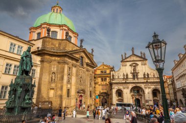 Akan bornozlu bir erkek heykeli, bir asa tutuyor ve el kol hareketi yapıyor, bir kamu meydanında bir sütun üzerinde duruyor ve arka planda bir köprü ve şehir manzarası var..