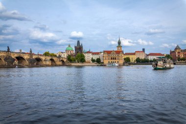 Tarihi bir köprü, bulutlu bir gökyüzü altında çeşitli mimari tarzlara sahip bir şehir manzarasıyla bir Avrupa şehrinde geniş bir nehir boyunca uzanır..